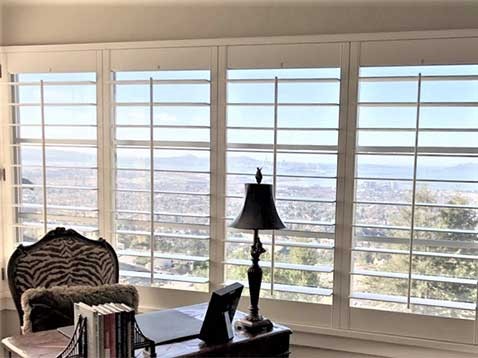 A desk with a lamp next to four large windows with shutters